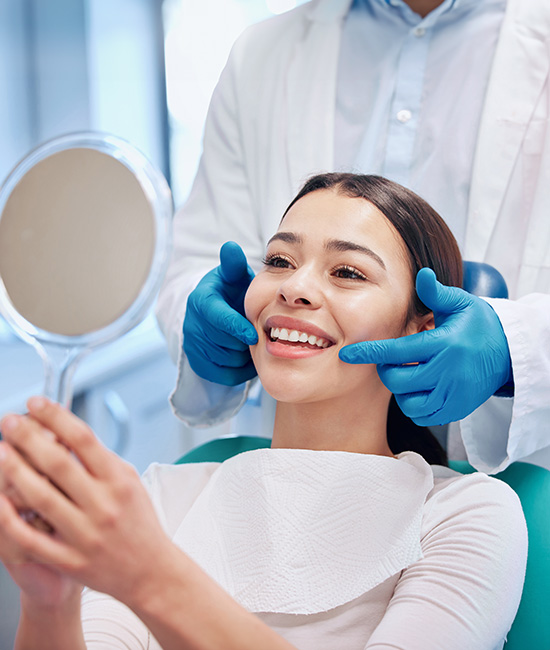 Woman sitting in dental chair with hygienist applying a face mask, both wearing blue gloves and green protective visors.